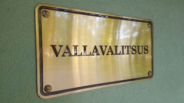 Pildil on lähedalt vaadatud metallist silt, millel on kiri „VALLAVALITSUS“. Silt on kuldset tooni ja läikiva pinnaga, millelt peegelduvad ümbritsevad puud või valgus. Tähed on tumedamad ja reljeefsed, andes sildile piduliku ja ametliku ilme. Silt on kinnitatud seinale nelja kruviga, üks igas nurgas. Taustaks on hele rohekas krohvitud sein, mis kontrastina toob kuldse sildi hästi esile. Üldmulje on ametlik ja esinduslik, viidates avaliku asutuse sissepääsule või hoonele.