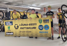 Lõuna-Eesti Rattaklubi avas Ülenurmel rattabaasi Treener koos noorrattasportlastega, kellest üks hoiab ratta nokka õhus. Esiplaanil toetajate bänner, taamal spordisaal ja jalgrattad.