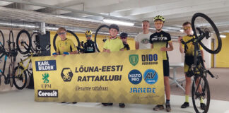 Lõuna-Eesti Rattaklubi avas Ülenurmel rattabaasi Treener koos noorrattasportlastega, kellest üks hoiab ratta nokka õhus. Esiplaanil toetajate bänner, taamal spordisaal ja jalgrattad.