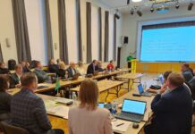 Vallavolikogu võttis vastu lisaeelarve ja kinnitas komisjonide koosseisud Pildil on kujutatud Kambja vallavolikogu istung Reola kultuurimajas. Suures saalis on U-kujuliselt paigutatud lauad, mille ümber istuvad vallavolikogu liikmed ja ametnikud. Enamik osalejatest jälgib ettekannet, osa töötab sülearvutite ja dokumentidega. Laudadel on näha sülearvuteid, pabermaterjale, kirjutusvahendeid ja veepudeleid. Saali eesosas seisab kõnepuldil ettekandja, kelle selja taga on suur ekraan. Ekraanil kuvatakse slaid pealkirjaga „2025. a III lisaeelarve“, kus on esitatud peamised eelarvenäitajad, sealhulgas lisaeelarve maht, eelarve kogumaht ning põhitegevuse tulude ja kulude muutused. Saali valgustavad laelambid ja lavavalgustus, ruum on kõrge ning heleda interjööriga. Üldmulje on ametlik ja töine – tegemist on kohaliku omavalitsuse otsustusprotsessi kajastava hetkega, kus arutatakse valla eelarve ja muid olulisi küsimusi.