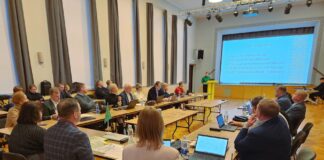 Vallavolikogu võttis vastu lisaeelarve ja kinnitas komisjonide koosseisud Pildil on kujutatud Kambja vallavolikogu istung Reola kultuurimajas. Suures saalis on U-kujuliselt paigutatud lauad, mille ümber istuvad vallavolikogu liikmed ja ametnikud. Enamik osalejatest jälgib ettekannet, osa töötab sülearvutite ja dokumentidega. Laudadel on näha sülearvuteid, pabermaterjale, kirjutusvahendeid ja veepudeleid. Saali eesosas seisab kõnepuldil ettekandja, kelle selja taga on suur ekraan. Ekraanil kuvatakse slaid pealkirjaga „2025. a III lisaeelarve“, kus on esitatud peamised eelarvenäitajad, sealhulgas lisaeelarve maht, eelarve kogumaht ning põhitegevuse tulude ja kulude muutused. Saali valgustavad laelambid ja lavavalgustus, ruum on kõrge ning heleda interjööriga. Üldmulje on ametlik ja töine – tegemist on kohaliku omavalitsuse otsustusprotsessi kajastava hetkega, kus arutatakse valla eelarve ja muid olulisi küsimusi.