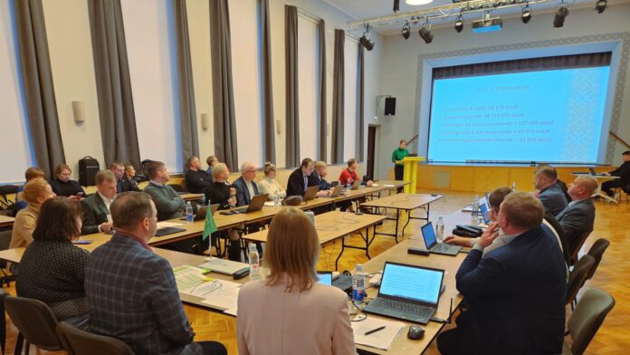 Pildil on kujutatud Kambja vallavolikogu istung Reola kultuurimajas. Suures saalis on U-kujuliselt paigutatud lauad, mille ümber istuvad vallavolikogu liikmed ja ametnikud. Enamik osalejatest jälgib ettekannet, osa töötab sülearvutite ja dokumentidega. Laudadel on näha sülearvuteid, pabermaterjale, kirjutusvahendeid ja veepudeleid. Saali eesosas seisab kõnepuldil ettekandja, kelle selja taga on suur ekraan. Ekraanil kuvatakse slaid pealkirjaga „2025. a III lisaeelarve“, kus on esitatud peamised eelarvenäitajad, sealhulgas lisaeelarve maht, eelarve kogumaht ning põhitegevuse tulude ja kulude muutused. Saali valgustavad laelambid ja lavavalgustus, ruum on kõrge ning heleda interjööriga. Üldmulje on ametlik ja töine – tegemist on kohaliku omavalitsuse otsustusprotsessi kajastava hetkega, kus arutatakse valla eelarve ja muid olulisi küsimusi.