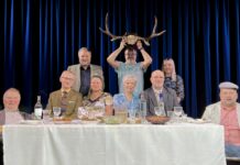 Kambja Püünelind on lavasid vallutanud juba 30 aastat Lavastseen: üheksa inimest istuvad ja seisavad kaetud laua ümber sinise eesriide taustal; keskel hoiab mees naljatamisi põdrasarvi pea kohal.