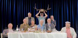 Kambja Püünelind on lavasid vallutanud juba 30 aastat Lavastseen: üheksa inimest istuvad ja seisavad kaetud laua ümber sinise eesriide taustal; keskel hoiab mees naljatamisi põdrasarvi pea kohal.