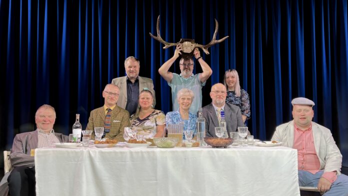 319_14_pyynelind_w Lavastseen: üheksa inimest istuvad ja seisavad kaetud laua ümber sinise eesriide taustal; keskel hoiab mees naljatamisi põdrasarvi pea kohal.