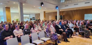 Esimesel alushariduse konverentsil peatuti kasvamisrõõmul Pildil on suur hulk inimesi kogunenud konverentsile või seminarile avarasse ja modernsesse saali. Osalejad istuvad ridades toolidel, enamik neist on suunatud ühele poole, ilmselt kuulamas esinejat, kes jääb kaadrist välja. Esireas on näha erinevas vanuses inimesi, riietatud pigem pidulikult või äristiilis. Paljudel on kaasas märkmikud või kotid, mõnedel rinnas nimekaardid, mis viitab ametlikule üritusele. Näoilmed on keskendunud ja tähelepanelikud. Ruum ise on hele ja kaasaegne: puidust seinapaneelid, kõrge lagi ning ühtlane valgustus loovad professionaalse õhkkonna. Laes on näha ka valgus- ja tehnikaseadmeid. Tagapool paistab avatud ala, mis võib olla kohvinurk või puhkeala. Üldmulje on organiseeritud ja rahulik, rõhutades õppimisele või arutelule suunatud sündmust.