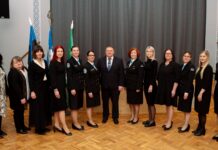 Kambjas loodi taas Naiskodukaitse jaoskond – sajandipikkune side mineviku ja tulevikuga Pildil on rühmaportree pidulikus ja ametlikus keskkonnas. Ühes reas seisab rühm naisi ning keskel üks mees, kes on riietatud ülikonda ja lipsu. Naised kannavad valdavalt musta värvi rõivaid; mitmel neist on naiskodukaitse vormiriietus, millel on nähtavad märgid ja embleemid. Taustal on näha Eesti lipp, naiskodukaitse lipp ja Kambja valla lipp, mis viitavad ametlikule või riikliku tähendusega sündmusele. Ruum on avar, kõrge laega ning parkettpõrandaga, andes pildile esindusliku ja piduliku ilme. Inimesed seisavad sirgelt ja vaatavad kaamerasse, nende ilmed on rahulikud ja väärikad. Üldmulje on organiseeritud ja tseremoniaalne, viidates näiteks tunnustamisüritusele, ametlikule koosviibimisele või organisatsiooni tähtpäevale.