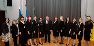 Kambjas loodi taas Naiskodukaitse jaoskond – sajandipikkune side mineviku ja tulevikuga Pildil on rühmaportree pidulikus ja ametlikus keskkonnas. Ühes reas seisab rühm naisi ning keskel üks mees, kes on riietatud ülikonda ja lipsu. Naised kannavad valdavalt musta värvi rõivaid; mitmel neist on naiskodukaitse vormiriietus, millel on nähtavad märgid ja embleemid. Taustal on näha Eesti lipp, naiskodukaitse lipp ja Kambja valla lipp, mis viitavad ametlikule või riikliku tähendusega sündmusele. Ruum on avar, kõrge laega ning parkettpõrandaga, andes pildile esindusliku ja piduliku ilme. Inimesed seisavad sirgelt ja vaatavad kaamerasse, nende ilmed on rahulikud ja väärikad. Üldmulje on organiseeritud ja tseremoniaalne, viidates näiteks tunnustamisüritusele, ametlikule koosviibimisele või organisatsiooni tähtpäevale.