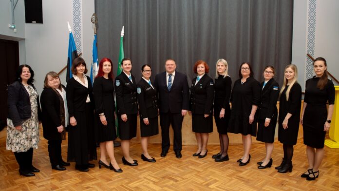 Pildil on rühmaportree pidulikus ja ametlikus keskkonnas. Ühes reas seisab rühm naisi ning keskel üks mees, kes on riietatud ülikonda ja lipsu. Naised kannavad valdavalt musta värvi rõivaid; mitmel neist on naiskodukaitse vormiriietus, millel on nähtavad märgid ja embleemid. Taustal on näha Eesti lipp, naiskodukaitse lipp ja Kambja valla lipp, mis viitavad ametlikule või riikliku tähendusega sündmusele. Ruum on avar, kõrge laega ning parkettpõrandaga, andes pildile esindusliku ja piduliku ilme. Inimesed seisavad sirgelt ja vaatavad kaamerasse, nende ilmed on rahulikud ja väärikad. Üldmulje on organiseeritud ja tseremoniaalne, viidates näiteks tunnustamisüritusele, ametlikule koosviibimisele või organisatsiooni tähtpäevale.