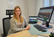 Saame tuttavaks Pildil on kujutatud noor naine istumas töölaua taga kontorikeskkonnas. Ta istub mugavas kontoritoolis, käed rahulikult laual, ning vaatab otse kaamerasse sõbraliku ja enesekindla ilmega. Tema ees laual on arvutitööks vajalikud vahendid: klaviatuur, hiir ja suur monitor, millel on avatud veebileht. Monitori kõrval on ka sülearvuti, mille ekraanil on sama või sarnane sisu. Laual on märkmepaberid, kirjutusvahendid ning kalender, mis viitab organiseeritud töökorraldusele. Taustal on hele sein ja seinal rippuv kalender. Paremal pool paistab aken, mille ees on õhuline kardin ning aknalaual toataim, mis lisab ruumile hubasust. Üldmulje on korrastatud ja rahulik töökeskkond.