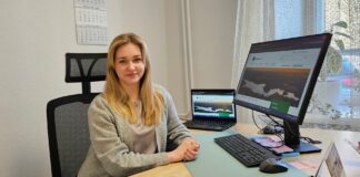 Saame tuttavaks Pildil on kujutatud noor naine istumas töölaua taga kontorikeskkonnas. Ta istub mugavas kontoritoolis, käed rahulikult laual, ning vaatab otse kaamerasse sõbraliku ja enesekindla ilmega. Tema ees laual on arvutitööks vajalikud vahendid: klaviatuur, hiir ja suur monitor, millel on avatud veebileht. Monitori kõrval on ka sülearvuti, mille ekraanil on sama või sarnane sisu. Laual on märkmepaberid, kirjutusvahendid ning kalender, mis viitab organiseeritud töökorraldusele. Taustal on hele sein ja seinal rippuv kalender. Paremal pool paistab aken, mille ees on õhuline kardin ning aknalaual toataim, mis lisab ruumile hubasust. Üldmulje on korrastatud ja rahulik töökeskkond.