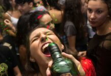 Alaealiste kodupeod ja riskikäitumine: politsei hoiatab sagenenud juhtumite eest Pildil on kujutatud rahvarohket pidu või ööklubi, kus noored inimesed tantsivad ja lõbutsevad värviliste valgusefektide keskel. Esiplaanil on noor naine, kelle pea on tahapoole kallutatud ja suu avatud, samal ajal kui keegi hoiab tema suu juures pudelit, justkui ta jooks sellest. Tema näoilme on intensiivne ja emotsionaalne, viidates tugevale hetkeelamusele. Taustal on tihedalt koos mitmeid teisi noori, kes räägivad, tantsivad või jälgivad toimuvat. Ühel paremal pool oleval neiul on pilk suunatud esiplaanil toimuvale, tema ilme on tõsisem ja tähelepanelik. Valgustus on hämar, kuid ruumi läbistavad rohelised ja punased laserilaadsed valguspunktid, mis loovad tüüpilise peokeskkonna atmosfääri. Kokkuvõttes annab pilt edasi elava ja veidi kaootilise peomeeleolu, kus tähelepanu keskmes on spontaanne ja intensiivne hetk.