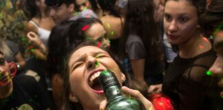 Alaealiste kodupeod ja riskikäitumine: politsei hoiatab sagenenud juhtumite eest Pildil on kujutatud rahvarohket pidu või ööklubi, kus noored inimesed tantsivad ja lõbutsevad värviliste valgusefektide keskel. Esiplaanil on noor naine, kelle pea on tahapoole kallutatud ja suu avatud, samal ajal kui keegi hoiab tema suu juures pudelit, justkui ta jooks sellest. Tema näoilme on intensiivne ja emotsionaalne, viidates tugevale hetkeelamusele. Taustal on tihedalt koos mitmeid teisi noori, kes räägivad, tantsivad või jälgivad toimuvat. Ühel paremal pool oleval neiul on pilk suunatud esiplaanil toimuvale, tema ilme on tõsisem ja tähelepanelik. Valgustus on hämar, kuid ruumi läbistavad rohelised ja punased laserilaadsed valguspunktid, mis loovad tüüpilise peokeskkonna atmosfääri. Kokkuvõttes annab pilt edasi elava ja veidi kaootilise peomeeleolu, kus tähelepanu keskmes on spontaanne ja intensiivne hetk.