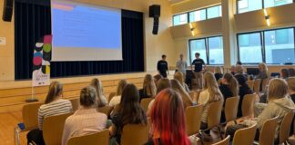 Kambja valla noorte karjääripäev kinnitas, et oma tee kujuneb tegutsedes Auditooriumis toimub esitlus, kus kolm noormeest tutvustavad lava ees seistes õppesuundi ja võimalusi. Ekraanil kuvatakse slaid „Õppekavad“, publikuks on peamiselt noored, kes kuulavad tähelepanelikult. Ruum on valgusküllane, suurte akendega ning istutakse ridades toolidel. Vasakul on näha stend kirjaga „LEEN“. Õhkkond on informatiivne ja suunatud hariduslike valikute tutvustamisele.