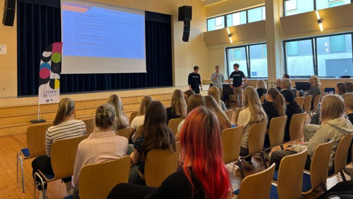 Auditooriumis toimub esitlus, kus kolm noormeest tutvustavad lava ees seistes õppesuundi ja võimalusi. Ekraanil kuvatakse slaid „Õppekavad“, publikuks on peamiselt noored, kes kuulavad tähelepanelikult. Ruum on valgusküllane, suurte akendega ning istutakse ridades toolidel. Vasakul on näha stend kirjaga „LEEN“. Õhkkond on informatiivne ja suunatud hariduslike valikute tutvustamisele.