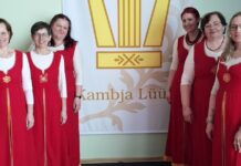 Kambja naisansambel Lüüra tegus märtsikuu Pildil poseerib kuus naist, kes kannavad ühtseid rahvuslikust stiilist inspireeritud punaseid kleite valgete pluusidega. Nad seisavad kahes grupis suure plakati kõrval, millel on kujutatud lüüra ning kiri „Kambja Lüüra“. Naised naeratavad ja vaatavad kaamerasse, luues sõbraliku ja ühtse meeleolu. Taustaks on hele sein ning põrandal puitparkett. Pilt viitab koorilaulu või kultuuritegevusega seotud kollektiivile.