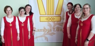 Kambja naisansambel Lüüra tegus märtsikuu Pildil poseerib kuus naist, kes kannavad ühtseid rahvuslikust stiilist inspireeritud punaseid kleite valgete pluusidega. Nad seisavad kahes grupis suure plakati kõrval, millel on kujutatud lüüra ning kiri „Kambja Lüüra“. Naised naeratavad ja vaatavad kaamerasse, luues sõbraliku ja ühtse meeleolu. Taustaks on hele sein ning põrandal puitparkett. Pilt viitab koorilaulu või kultuuritegevusega seotud kollektiivile.