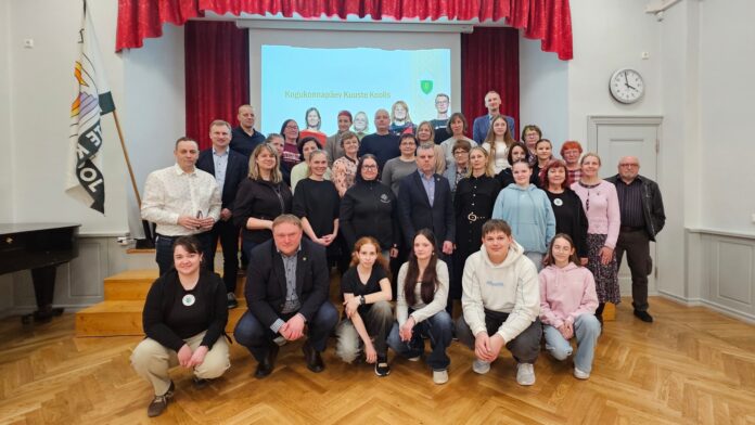 320_3_kogukonnapaev_kuustes Koolisaalis tehtud grupifotol poseerib suur hulk eri vanuses inimesi – õpilased, õpetajad ja kogukonna liikmed. Esireas istuvad noored, nende taga seisavad täiskasvanud. Taustal on lava punaste kardinate ja projektoriekraaniga, millel on kiri „Kogukonnapäev Kuuste koolis“. Saali seinal on kell ning vasakul pool seisab lipp. Õhkkond on pigem sõbralik, viidates ühisele sündmusele ja kogukonna koostööle.