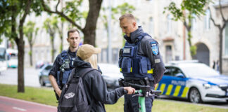 Elektritõukerataste kontroll näitab, et laste liikluskäitumine on murettekitav Linnatänaval peatavad kaks politseinikku elektritõukerattaga sõitva noore inimese. Vestlus toimub rahulikult: üks ametnikest seisab sõitja ees ja selgitab midagi, teine jälgib olukorda veidi eemal. Taustal on park, puud ja ajaloolised hooned ning tee ääres politseiauto. Tõukeratas on roheline ning sõitjal on seljas tume riietus ja seljakott. Stseen viitab liiklusjärelevalvele või ohutuskontrollile linnakeskkonnas.