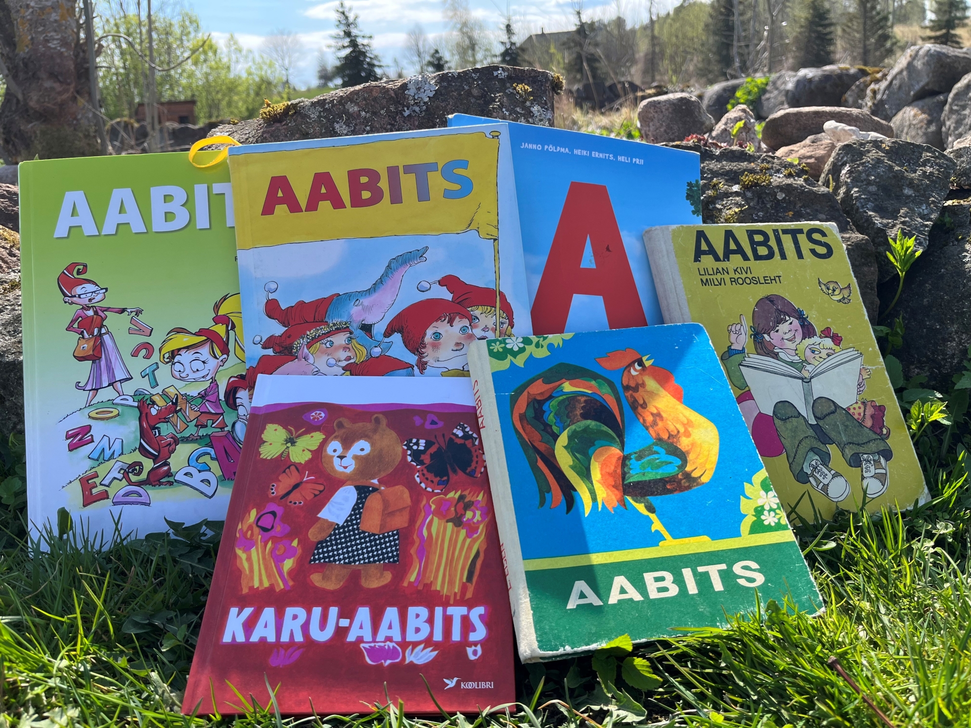 Murule ja aiakividele on seatud erinevad Eesti aabitsad, nende seas nn „Kuke aabits“ ja „Karu-aabits“. Taustal paistab kevadine loodus – rohelised puud, sinine taevas ja päikesevalgus.