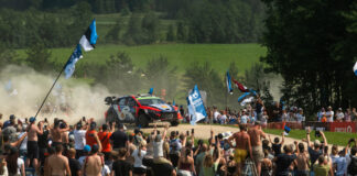 WRC Delfi Rally Estonial saab näha maailma parimaid ja noori Eesti tippsõitjaid Esiplaanil seisab tihe rahvahulk, kes jälgib kruusateel kihutavat ralliautot. Paljud inimesed hoiavad käes Eesti lippe ja meeskonnalippe ning filmivad või pildistavad sündmust telefonidega. Keskel sõidab värviline ralliauto suure tolmupilve saatel kurvi läbides. Rada on piiratud turvavõrkude ja reklaambänneritega. Taustal on näha rohelised põllud ja metsaservad, mis loovad suvise Lõuna-Eesti maastiku meeleolu