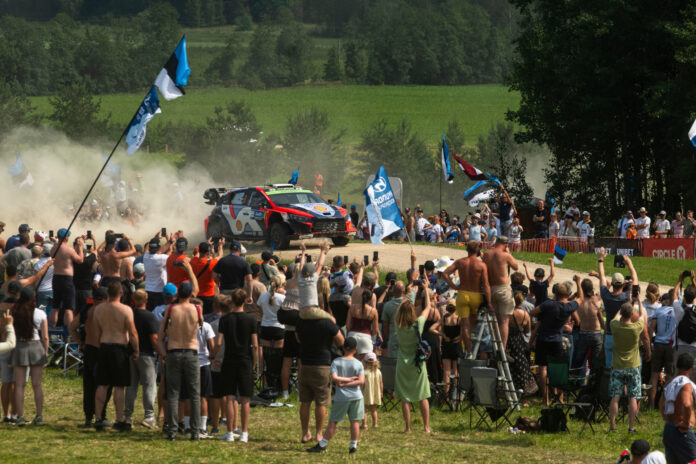 322_10_rally_estonia Esiplaanil seisab tihe rahvahulk, kes jälgib kruusateel kihutavat ralliautot. Paljud inimesed hoiavad käes Eesti lippe ja meeskonnalippe ning filmivad või pildistavad sündmust telefonidega. Keskel sõidab värviline ralliauto suure tolmupilve saatel kurvi läbides. Rada on piiratud turvavõrkude ja reklaambänneritega. Taustal on näha rohelised põllud ja metsaservad, mis loovad suvise Lõuna-Eesti maastiku meeleolu