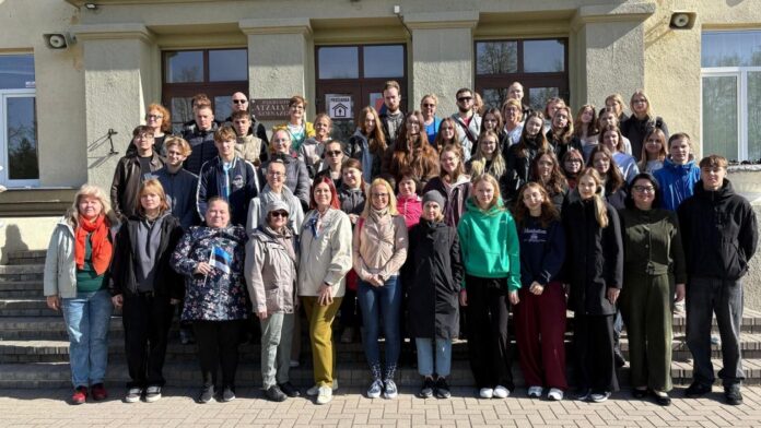 Suur rühm noori ja täiskasvanuid seisavad koolihoone ees trepil. Osalejad seisavad mitmes reas päikeselise ilmaga ning kannavad kevadisi jopesid ja vabaajariideid. Mõnel inimesel on käes Eesti lipp või seljakott. Taustal on näha koolimaja sissepääs ja hoone fassaad.