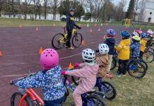 Kambja lasteaia tegemistest Laste rattakoolitus õues spordiväljakul. Väikesed lapsed seisavad või istuvad jalgratastel reas ning kannavad värvilisi kiivreid ja kevadriideid. Nende ees räägib juhendaja kollase jalgratta kõrval ning selgitab harjutust või liiklusreegleid. Väljakule on paigutatud oranžid koonused, mis moodustavad sõidurada või harjutusala. Taustal on näha puid, veekogu ja valge kirikuhoone.
