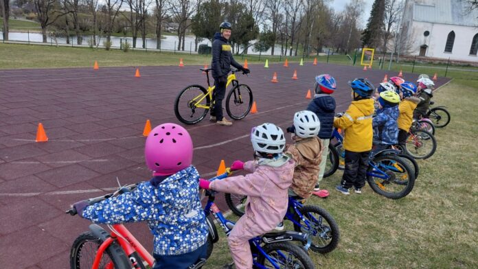 Rattakoolitus Kambja lasteaias Laste rattakoolitus õues spordiväljakul. Väikesed lapsed seisavad või istuvad jalgratastel reas ning kannavad värvilisi kiivreid ja kevadriideid. Nende ees räägib juhendaja kollase jalgratta kõrval ning selgitab harjutust või liiklusreegleid. Väljakule on paigutatud oranžid koonused, mis moodustavad sõidurada või harjutusala. Taustal on näha puid, veekogu ja valge kirikuhoone.