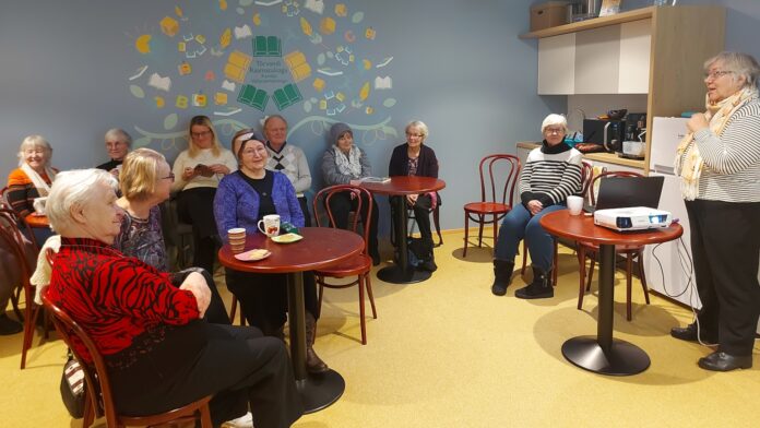 322_14_vanemaealiste_kogu Väikeste laudade ümber istuvad eakamad inimesed, kes kuulavad ruumi paremas servas kõnelevat naist. Laual on projektor ja sülearvuti, mis viitavad esitlusele või loengule. Osalejad istuvad hubases ja rahulikus õhkkonnas ning mitmel laual on tassid ja suupisted.