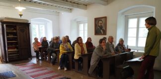 Ülenurme käsitööseltsi kosutav kultuurireis Mulgimaale Ekskursioonigrupp kuulab giidi ajaloolises koolitoas või muuseumiruumis. Külastajad istuvad vanades puidust koolipinkides ning ruumi ees räägib giid.