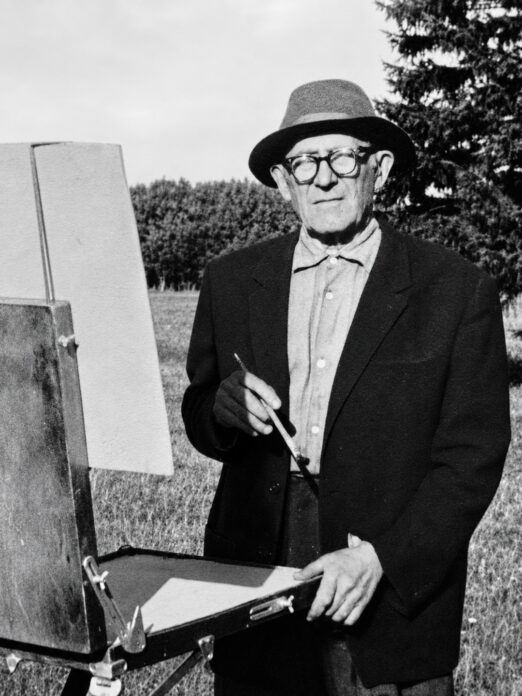 Mustvalgel fotol seisab eakas mees õues maalimisvarustuse kõrval. Tal on peas hele kaabu, ees prillid ning seljas tume pintsak heleda särgi peal. Mees hoiab käes pintslit ja seisab kokkupandava maalikasti või molberti juures, mille kõrval on tühi maalialus. Taustal paistab avatud rohumaa ning kõrged puud.